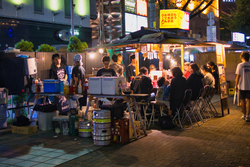 Yatai Streetfood-Stand bei Nacht in Fukuoka Ein beleuchteter Yatai-Straßenimbiss in Fukuoka bei Nacht, an dem Gäste auf Hockern sitzen und essen.