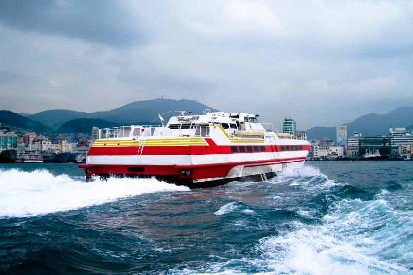 Schnellfähre auf dem Weg zwischen Fukuoka und Busan Eine rot-weiße Schnellfähre fährt bei leicht bewölktem Himmel über das Meer, im Hintergrund ist eine Küstenstadt mit Bergen zu sehen.