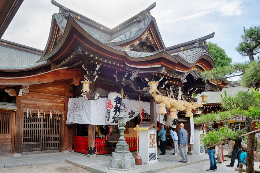 Der Kushida-Schrein in Fukuoka Das hölzerne Hauptgebäude des Kushida-Schreins in Fukuoka mit einem großen Strohseil (Shimenawa) und weißen Papierlaternen, davor beten Besucher.