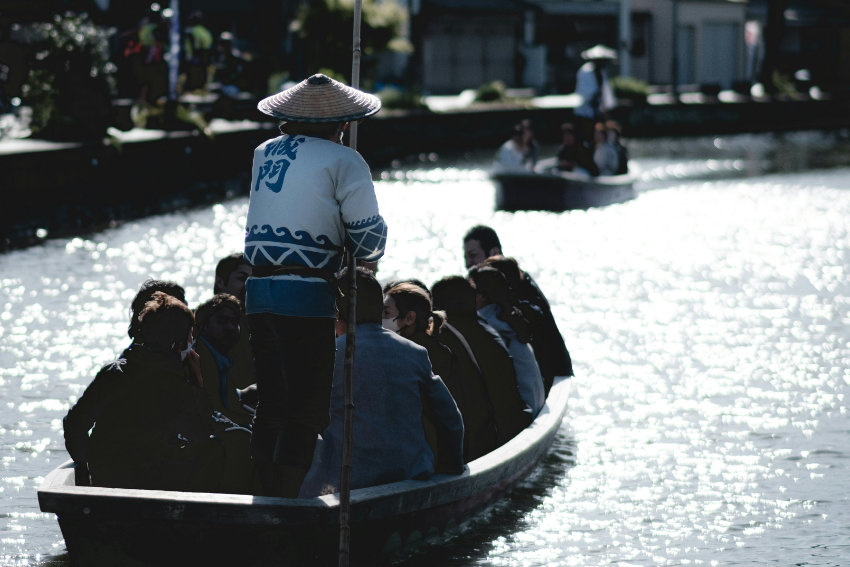 Bootsfahrt auf den Kanälen in Yanagawa Ein Bootsführer in traditioneller Kleidung und mit Strohhut steuert ein flaches Holzboot voller Passagiere durch einen sonnenbeschienenen Kanal in Yanagawa.