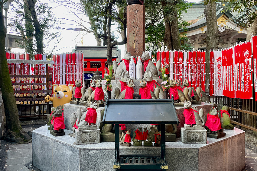 Toyokawa Inari Schrein in Tokio mit Kitsune-Statuen Steinfüchse mit roten Lätzchen am Toyokawa Inari Schrein in Tokio, umgeben von roten Fahnen und Opfergaben