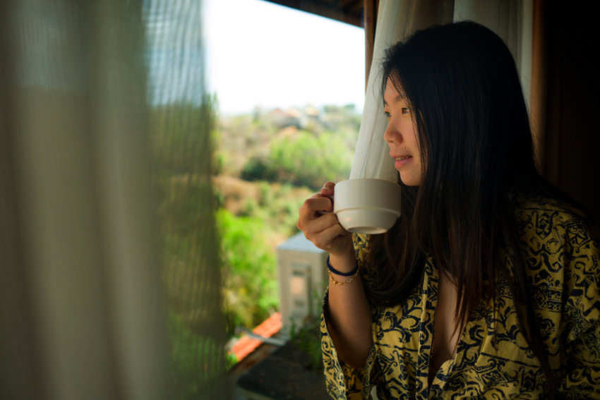Junge Frau blickt mit einer Tasse in der Hand gedankenverloren aus dem Fenster – ein Sinnbild für das japanische Wort Boketto
