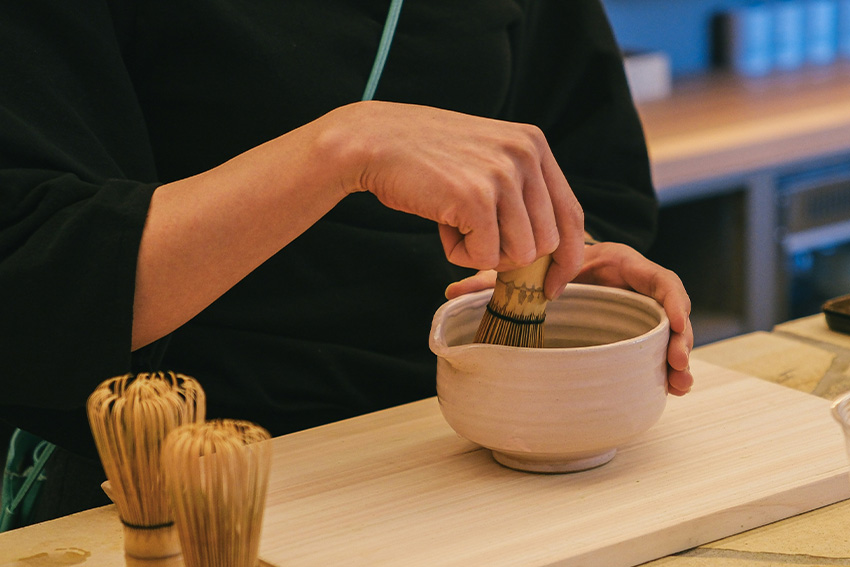 Hände bereiten Matcha mit einem Bambusbesen in einer Teeschale zu – Symbol für Sandō und den Übungsweg der Aufmerksamkeit in Japans drei Wegen (Sadō, Kadō, Kōdō).