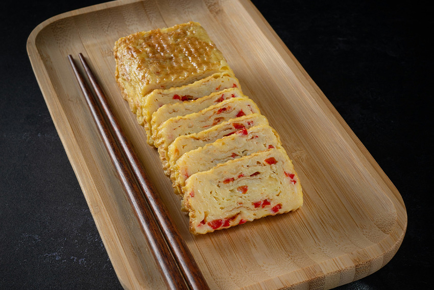 Tamagoyaki – japanisches Omelett aus gerolltem Ei mit Gemüse, in Scheiben geschnitten auf einem Holztablett mit Essstäbchen serviert