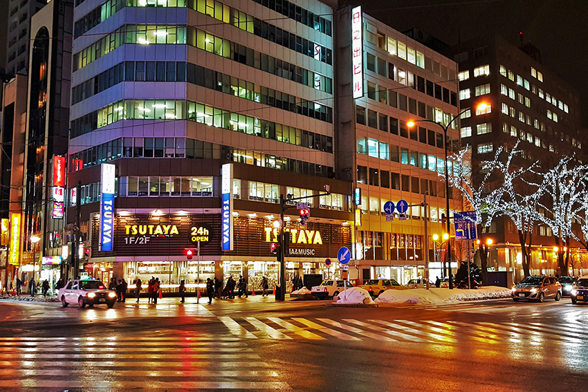 Belebte Kreuzung in Sapporo bei Nacht mit beleuchteten Gebäuden, winterlichen Straßen und urbaner Atmosphäre
